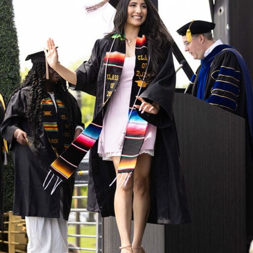 A student in commencement regalia and Hispanic affinity stole walks across a stage