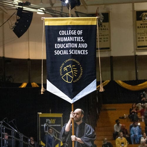 A graduate marches with a College of Humanities, Education and Social Sciences banner at PNW commencement