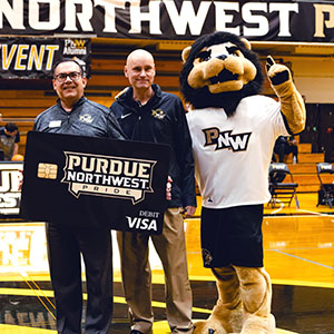 Two men hold up a mock Purdue University Northwest credit card beside PNW mascot Leo center court at a basketball game.