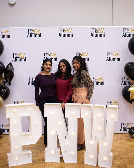 Three alumni stand together in front of a PNW alumni backdrop.