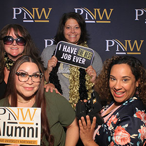 Four people pose in front of a PNW branded backdrop. They are all wearing or holding photo booth props