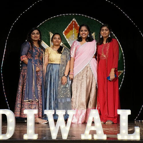 Four students in traditional Indian saris stand behind a marquee Diwali sign