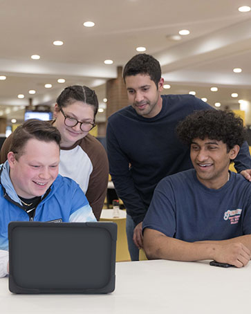 Four students look at a laptop. Two students are sitting at a table and two are standing behind them