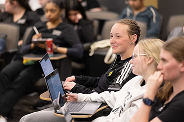 Students sit at desks in a classroom. They have laptops open in front of them