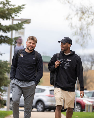 Two students walk together outside. There are trees and a parking lot behind them