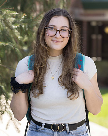 A student stands outside and smiles. They are wearing a beige shirt, glasses, and a backpack