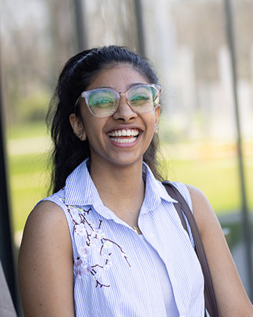 A student wears glasses and a purple shirt while standing outside. They are smiling.