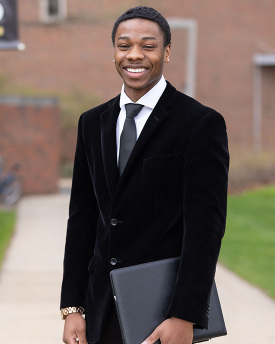 A student stands outdoors in a black suit and tie