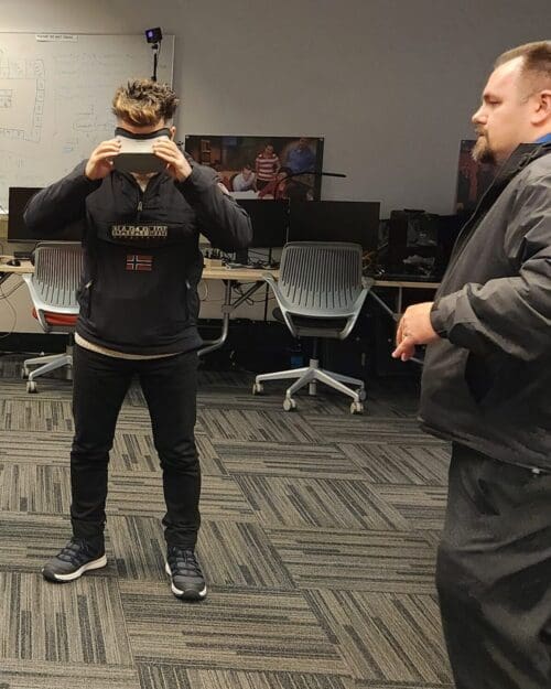 Visitor participates in hands-on experiences of AR & VR projects in the visualization lab. Participant is standing while holding VR goggles. Another individual gives instruction.