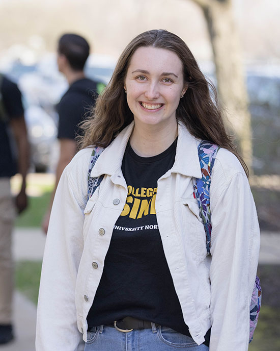 A student stands outside. They are wearing a white denim jacket and a black College of Business shirt