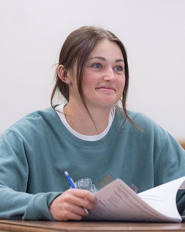 A student in a blue sweatshirt sits in a classroom. They are holding open a packet of papers