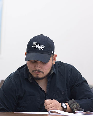 A student in a black shirt and black PNW hat sits at a desk and looks down at a piece of paper