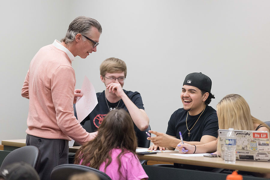 Students sit at a desk and laugh. They are talking with a professor