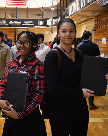 Two students stand back to back and hold up portfolios during a career expo