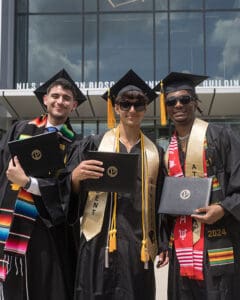 Three students stand in commencement regalia and hold up their diplomas