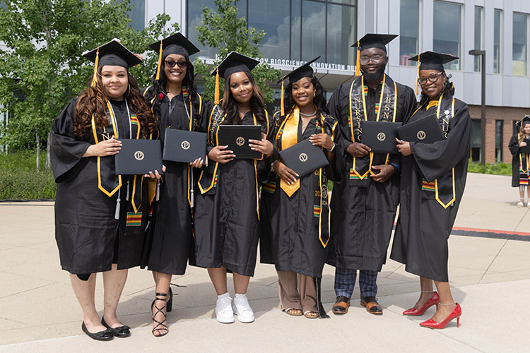 Six students pose together in commencement regalia and hold up their diplomas