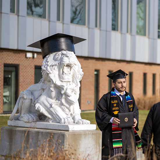 A PNW graduate poses in front of a lion sculpture in commencement regalia.