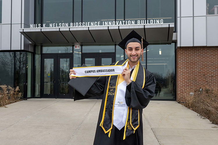 A PNW graduate holds up a "Campus Ambassador" sign in front of the Nils K. Nelson Bioscience Innovation Building.