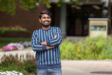 A student in a blue striped long sleeve shirt crosses their arms and smiles