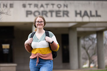 A student in a striped sweater holds their backpack straps. They are standing alone and smiling.
