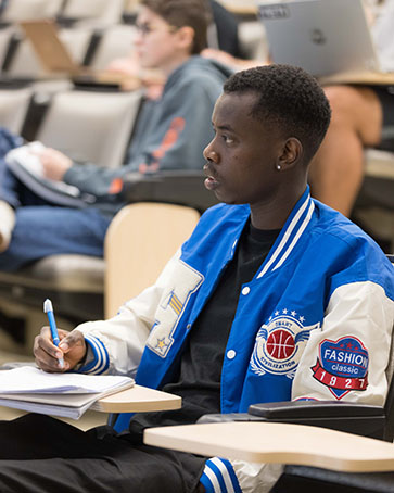 A student sits in a lecture hall. They are wearing a blue and white varsity jacket