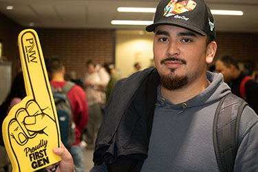 A student holds up a yellow "Proudly First-Gen" foam finger. They are wearing a hat and a gray hoodie