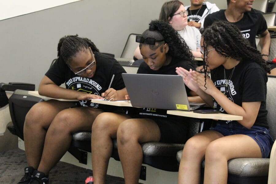 Three female students sitting next to each other. Student on the left is writing. Student in the middle is on the computer. Student on the right is talk to the other two, with her hands.