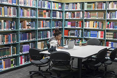 PNW student sitting at a table in library, surrounded by filled bookshelves and looking at laptop.
