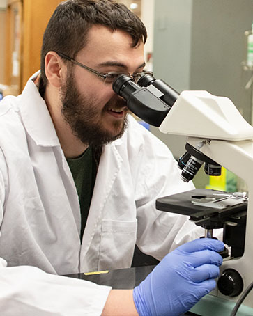 A student in a white lab coat and blue disposable gloves looks into a microscope