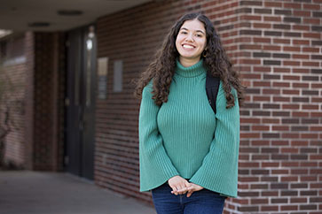 A student with dark curly hair wears a green turtleneck sweater and backpack. They are standing outside in front of a brick wall