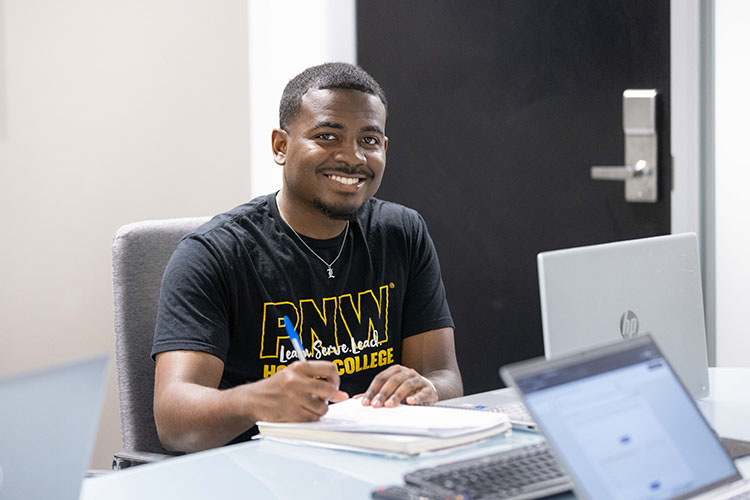 PNW Honors College student sitting at desk with pen in hand and notebook on table, smiling at camera.