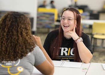 Two students sit at a table. There are notebooks open in front of them.