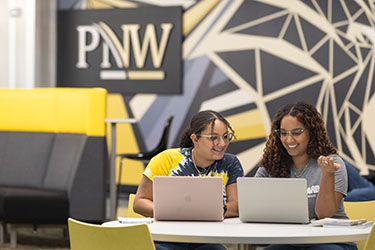 Two students sit at a table. They both have open laptops in front of them