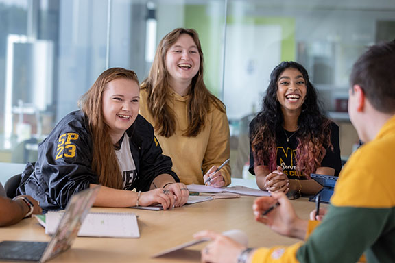 Four students sit at a table. Three of the students are facing the camera and smiling