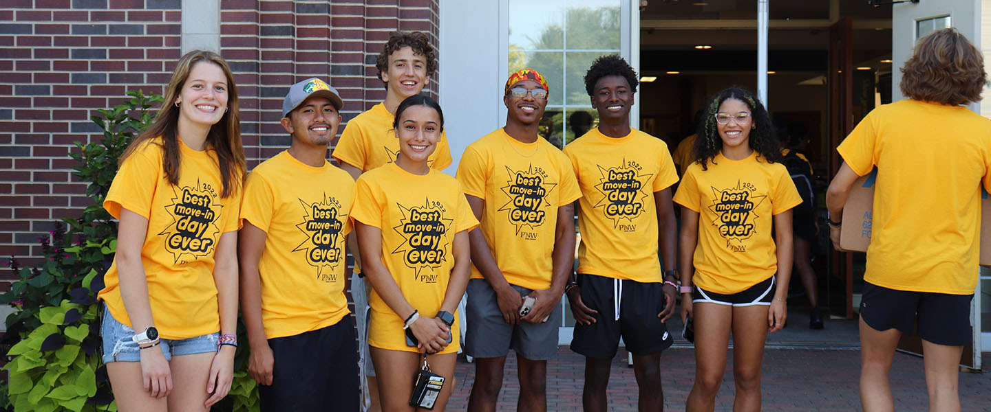 Eight students in yellow "Move-in Day" shirts stand together outside of one of the dorm buildings