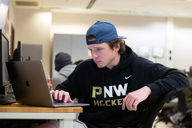 A student in a PNW Hockey hoodie works at a laptop.
