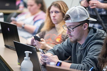 Students sit at a long table and look at their phones