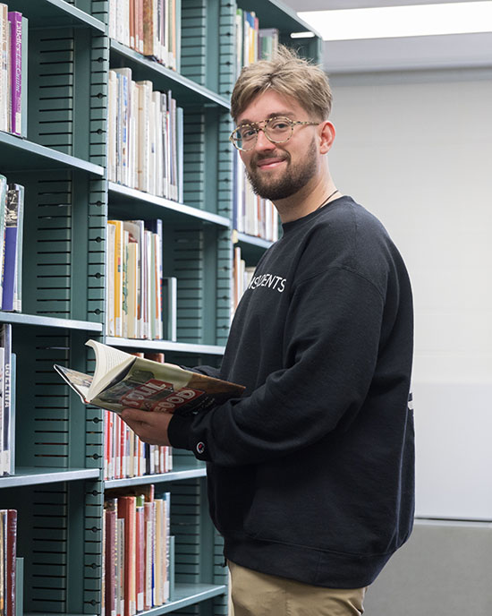 A student stands in front of a library bookshelf. They are holding a book open and smiling at the camera