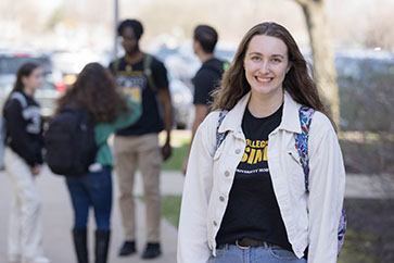 A student in a college of business t-shirt, white jean jacket and backpack stands alone and smiles. There are other students in the background.