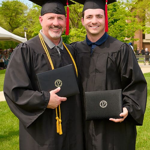 Jacob Wheeler Sr. and Jacob Wheeler Jr. stand together in commencement regalia. They are holding their diplomas.