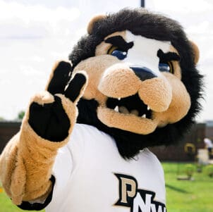 PNW lion mascot Leo raises two fingers at PNW's outdoor Dowling Park field.