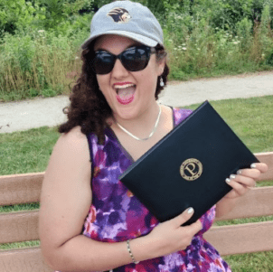 Katie Beverly, sitting on a bench in a park, smiling and holding her Purdue Diploma