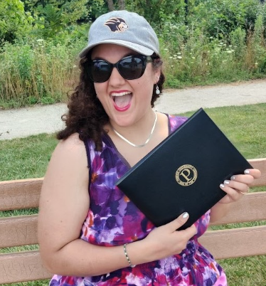 Katie Beverly, sitting on a bench in a park, smiling and holding her Purdue Diploma