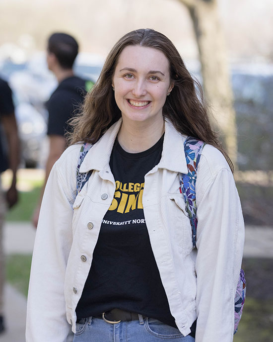 A student stands outdoors in a black College of Business t-shirt and white denim jacket.