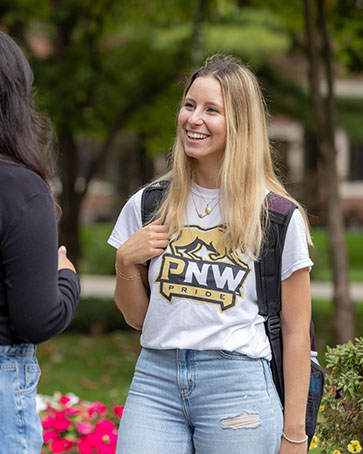 A student with long blonde hair stands in a white PNW Pride t-shirt and light blue jeans.