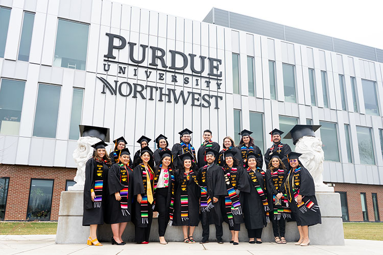 A group of PNW students pose for a picture in front of the NILS building in Hammond. The students are wearing graduation cap and gowns and honorary stoles.