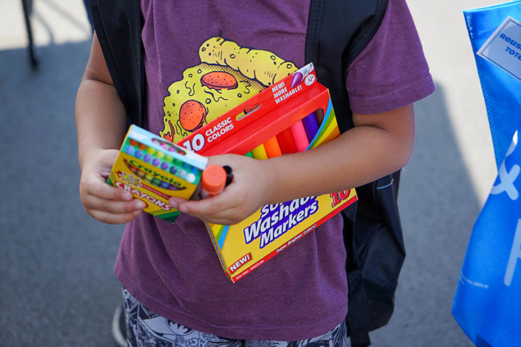 A child holds a glue stick, box of crayons and washable markers