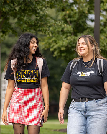 Two students walk outdoors on campus. The student on the left is wearing a PNW Honors shirt with a red skirt. The student on the right is wearing a Powering Onward shirt with blue jeans.