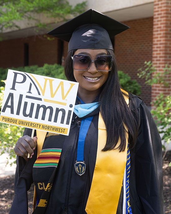 An alumni stands in commencement regalia and holds up a PNW Alumni sign