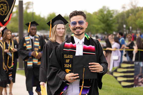 A Spring 2024 graduate in a Latino affinity stole and commencement regalia holds their cap in front of them and smiles